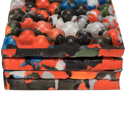 A close-up of a stack of thick, textured foam mats with a bold abstract paint-splatter pattern in orange, black, red, blue, green, and white, set against a plain white background.