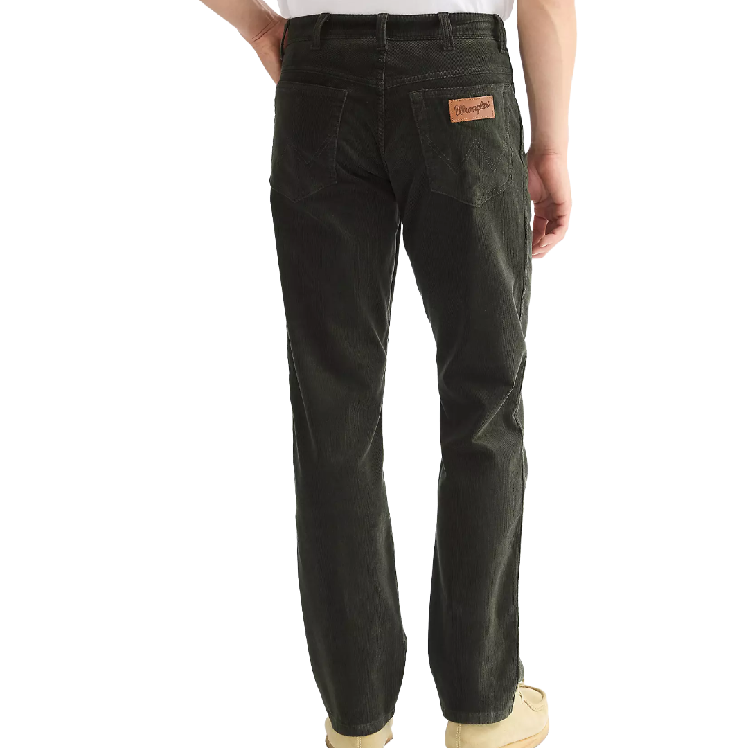 Rear view of a person wearing dark charcoal gray denim jeans with a brown leather Wrangler patch on the back pocket, paired with a white shirt and beige shoes, on a white background.