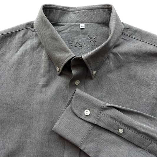 A close-up of a gray long-sleeve button-down shirt with a spread collar and folded cuff, showing the 'Burb Regent' brand name printed inside the collar.