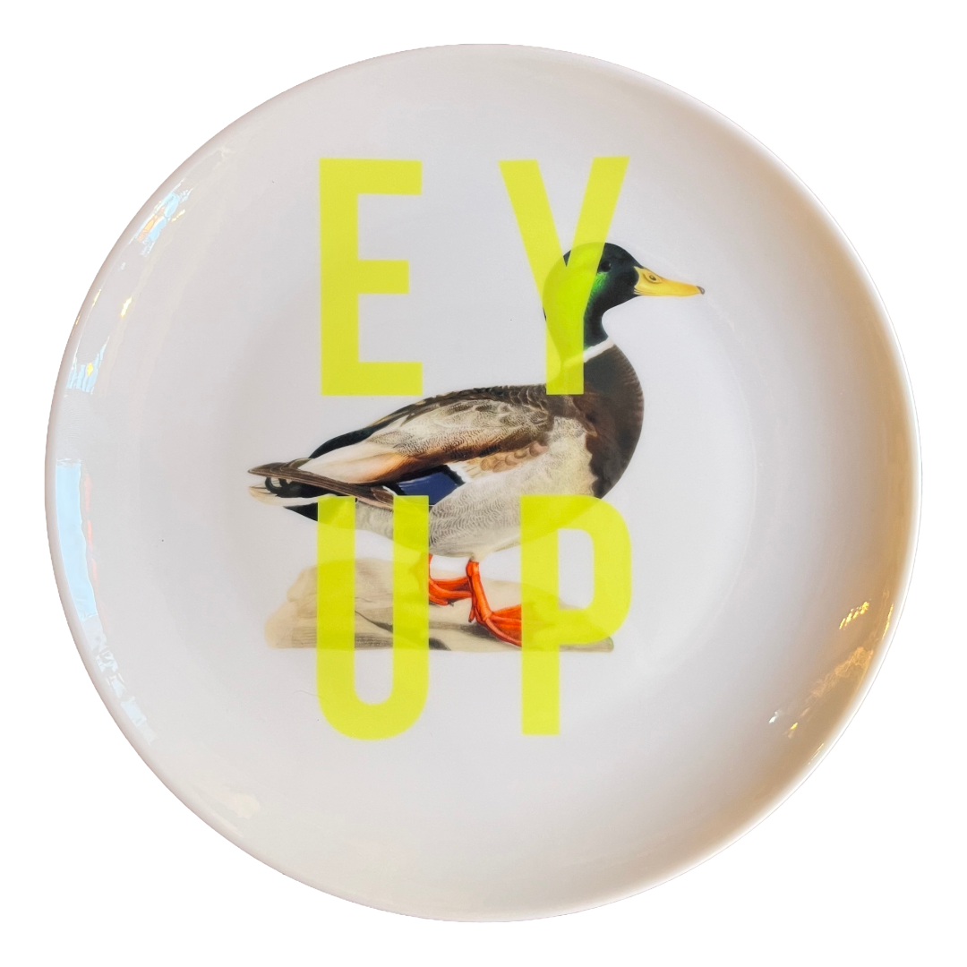 A white ceramic plate featuring a printed mallard duck graphic with large, bold yellow text overlay reading 'EY UP'.