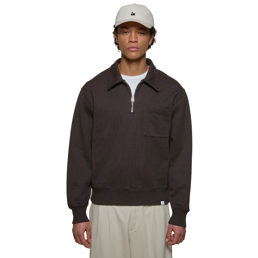 A model wearing a dark brown ribbed quarter-zip pullover with a collar and chest pocket, styled with a white t-shirt, beige pants, and a beige cap.