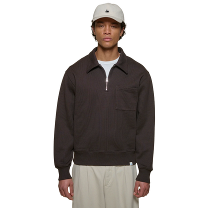 A model wearing a dark brown ribbed quarter-zip pullover with a collar and chest pocket, styled with a white t-shirt, beige pants, and a beige cap.