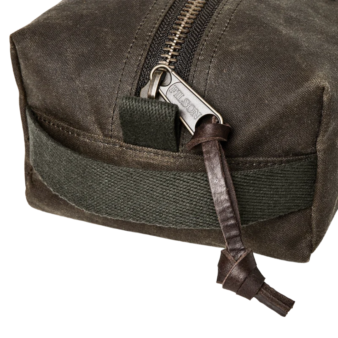 Close-up of a dark olive green waxed canvas pouch with a silver-toned metal zipper, a dark olive green webbing loop, and a brown leather zipper pull tab engraved with the brand name 'FALCON'.