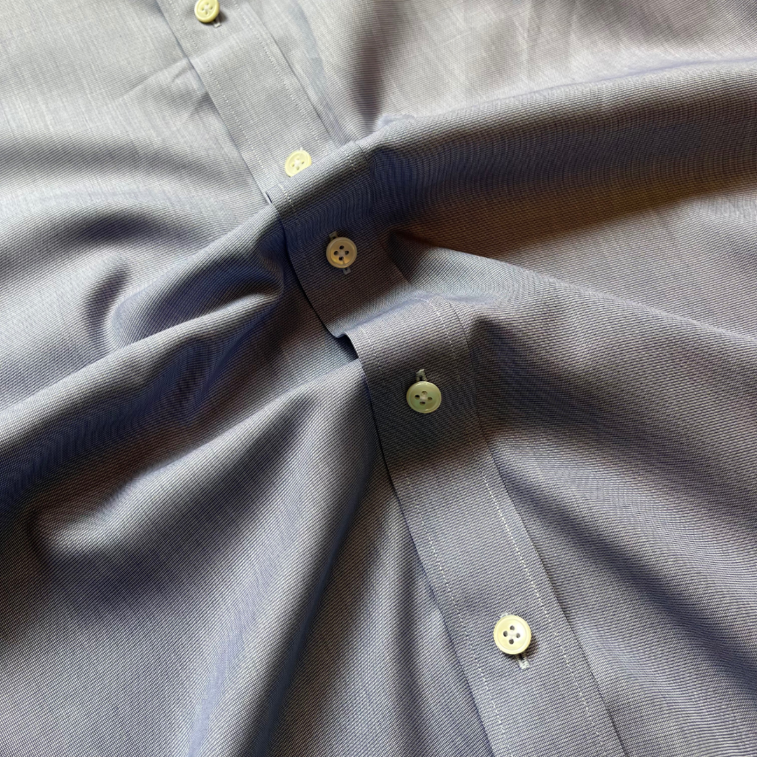 Close-up of a light blue-gray textured button-down shirt with pale yellow buttons, showing the fabric weave and collar construction.