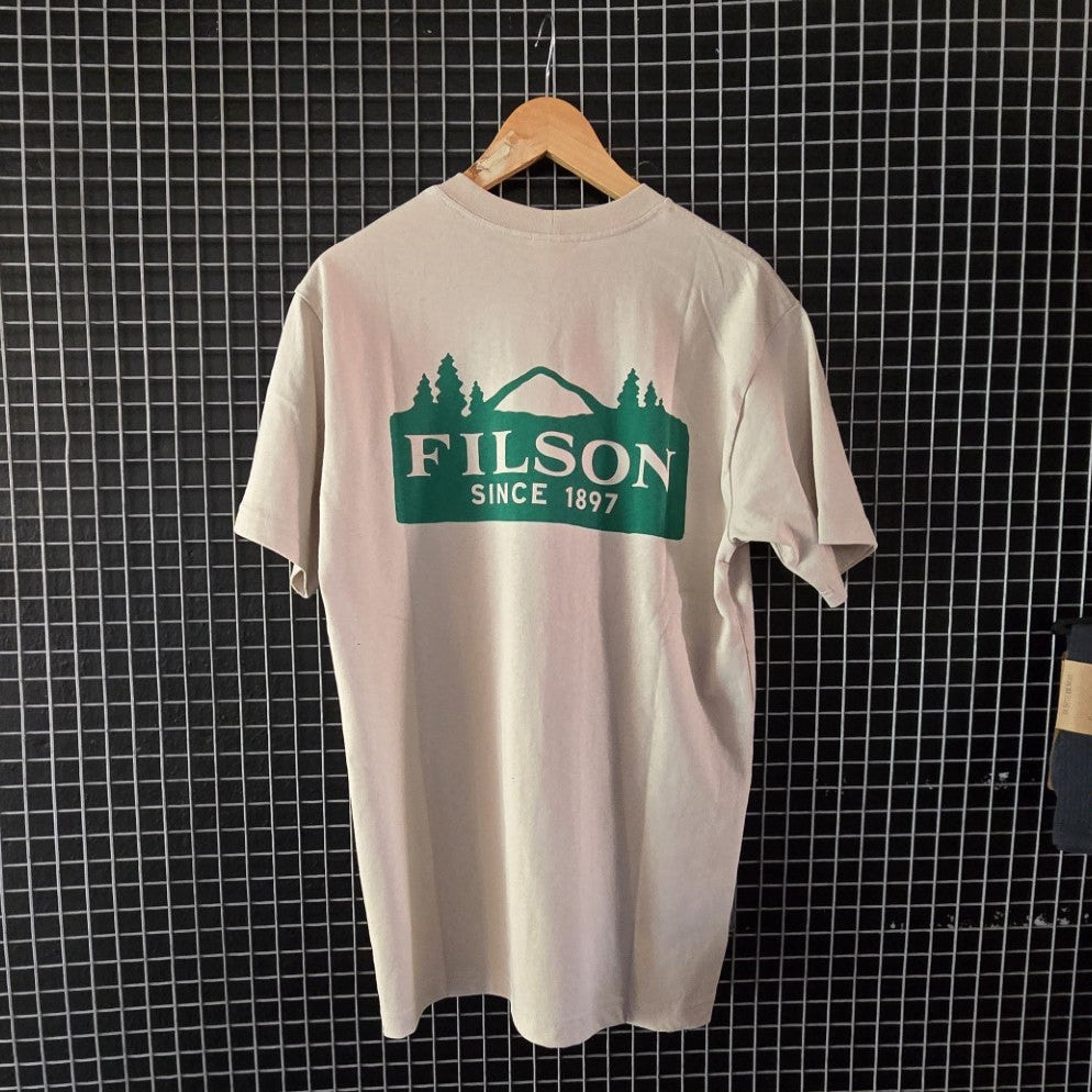 Beige t-shirt with Filson logo hanging on a grid wall with bags below.