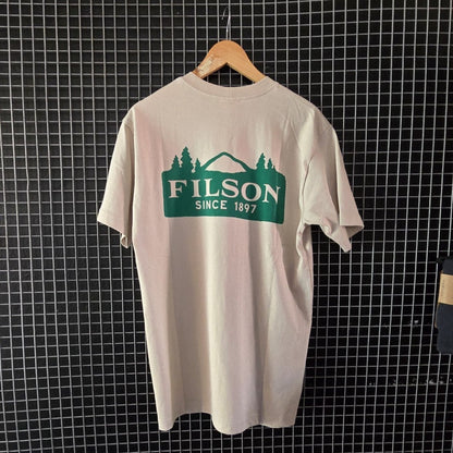 Beige t-shirt with Filson logo hanging on a grid wall with bags below.