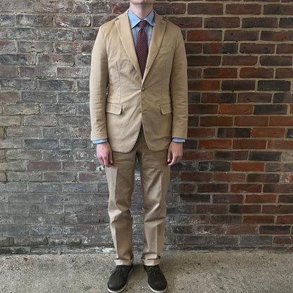 Man wearing a Regent 'George' beige cotton suit with a blue shirt and a burgundy tie with white polka dots standing against a brick wall