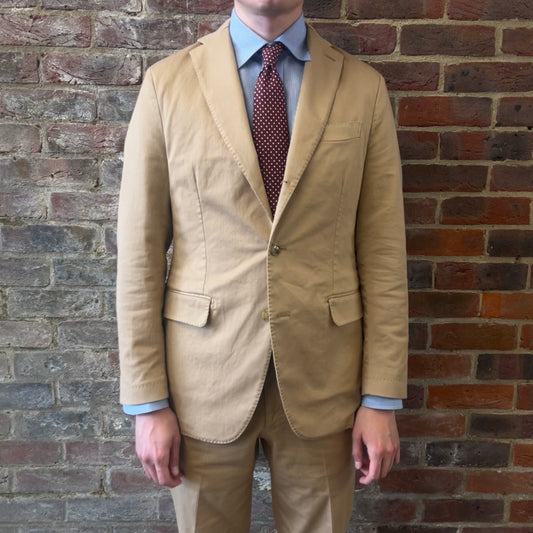Man wearing a Regent 'George' beige cotton suit with a blue shirt and a burgundy tie with white polka dots standing against a brick wall