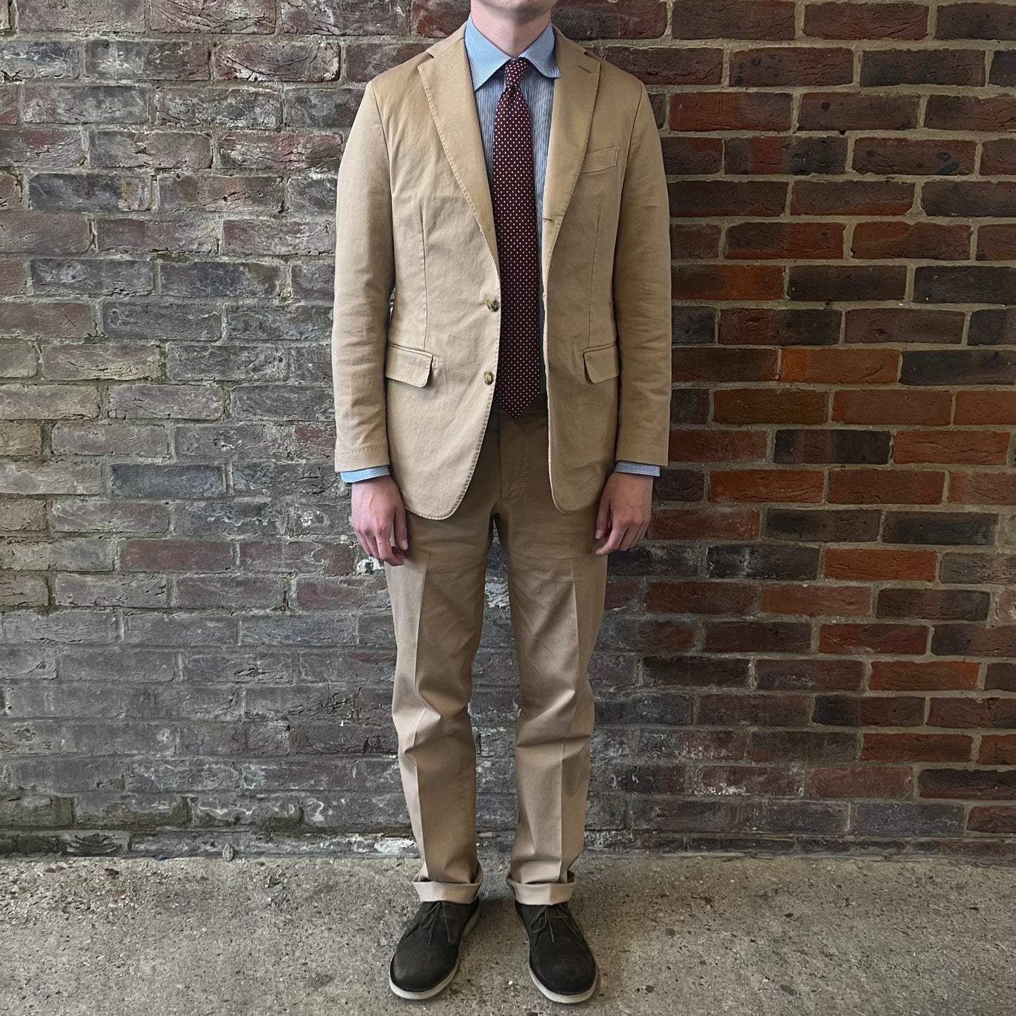 Man wearing a Regent 'George' beige cotton suit with a blue shirt and a burgundy tie with white polka dots standing against a brick wall