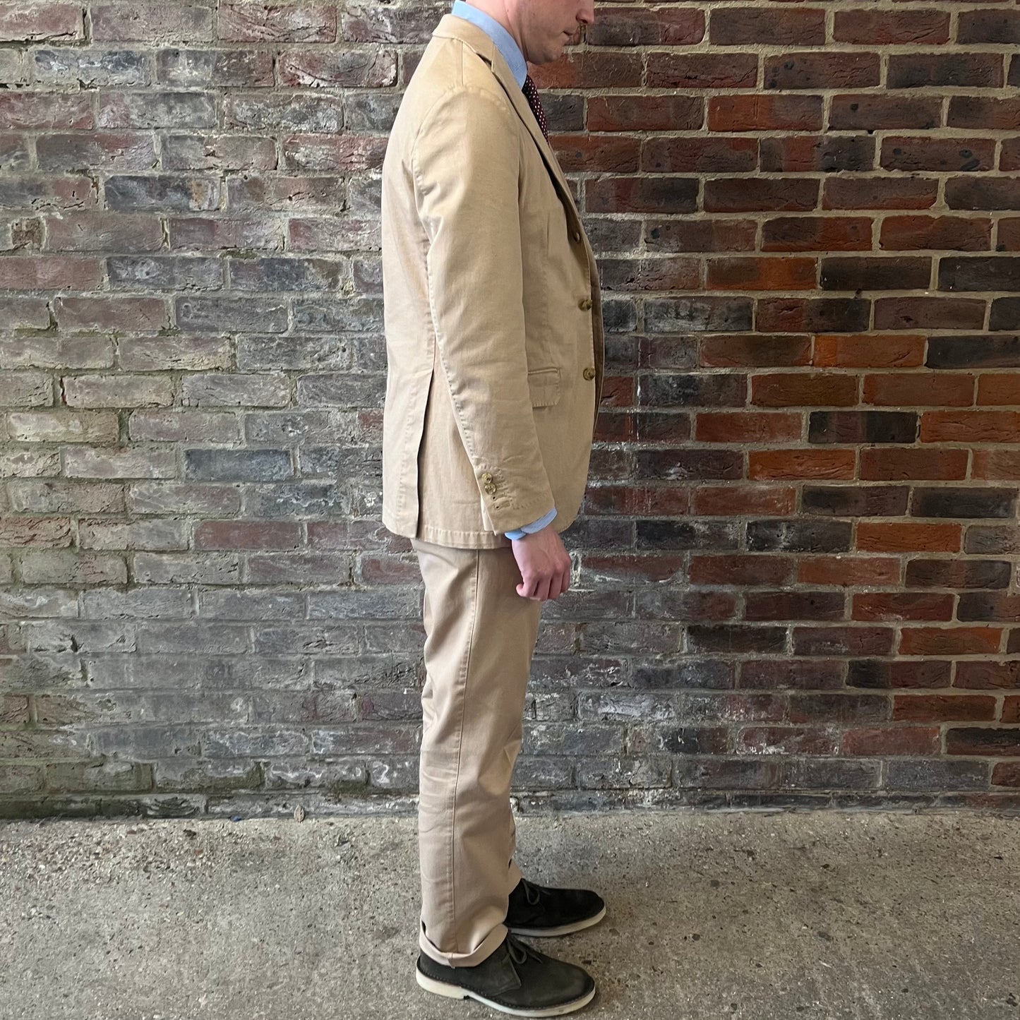 Man wearing a Regent 'George' beige cotton suit with a blue shirt and a burgundy tie with white polka dots standing against a brick wall