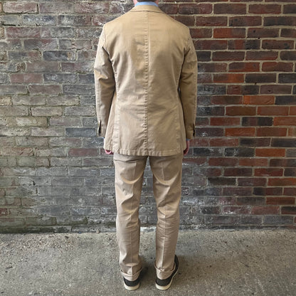 Man wearing a Regent 'George' beige cotton suit with a blue shirt and a burgundy tie with white polka dots standing against a brick wall facing the wall showing off the double stitch detail on the rear