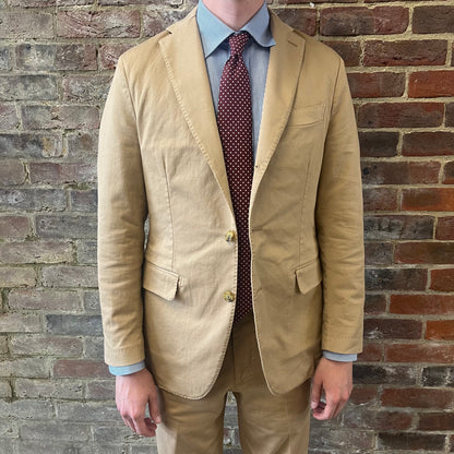 Man wearing a Regent 'George' beige cotton suit with a blue shirt and a burgundy tie with white polka dots standing against a brick wall