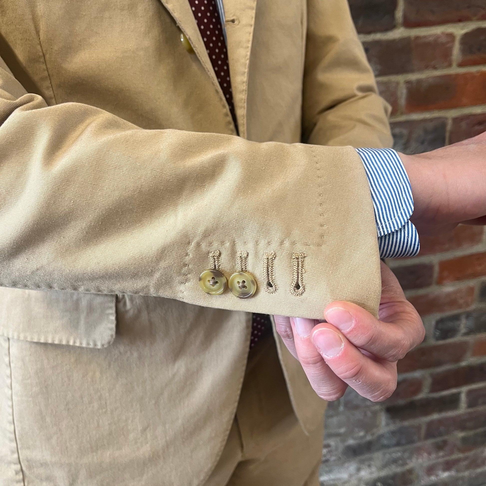 Man wearing a Regent 'George' beige cotton suit with a blue shirt and a burgundy tie with white polka dots standing against a brick wall showing off the jacket's working cuff detailing