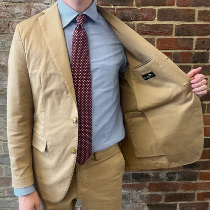 Man wearing a Regent 'George' beige cotton suit with a blue shirt and a burgundy tie with white polka dots standing against a brick wall showing off the lining of his jacket