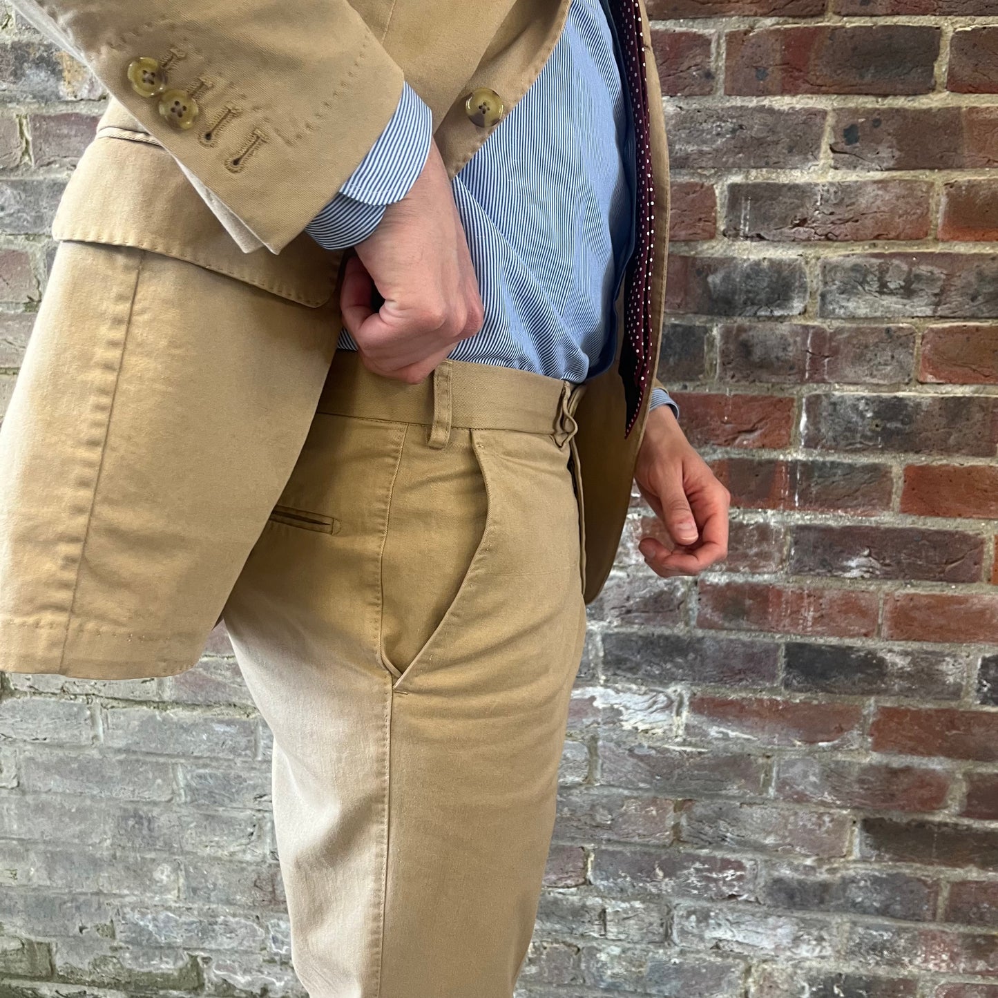 Man wearing a Regent 'George' beige cotton suit with a blue shirt and a burgundy tie with white polka dots standing against a brick wall demonstrating the belt loops on his trousers