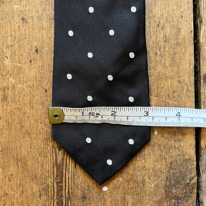 A black necktie with white polka dots laid flat on a rustic wooden surface, with a metal tape measure placed horizontally across the middle to show the size.
