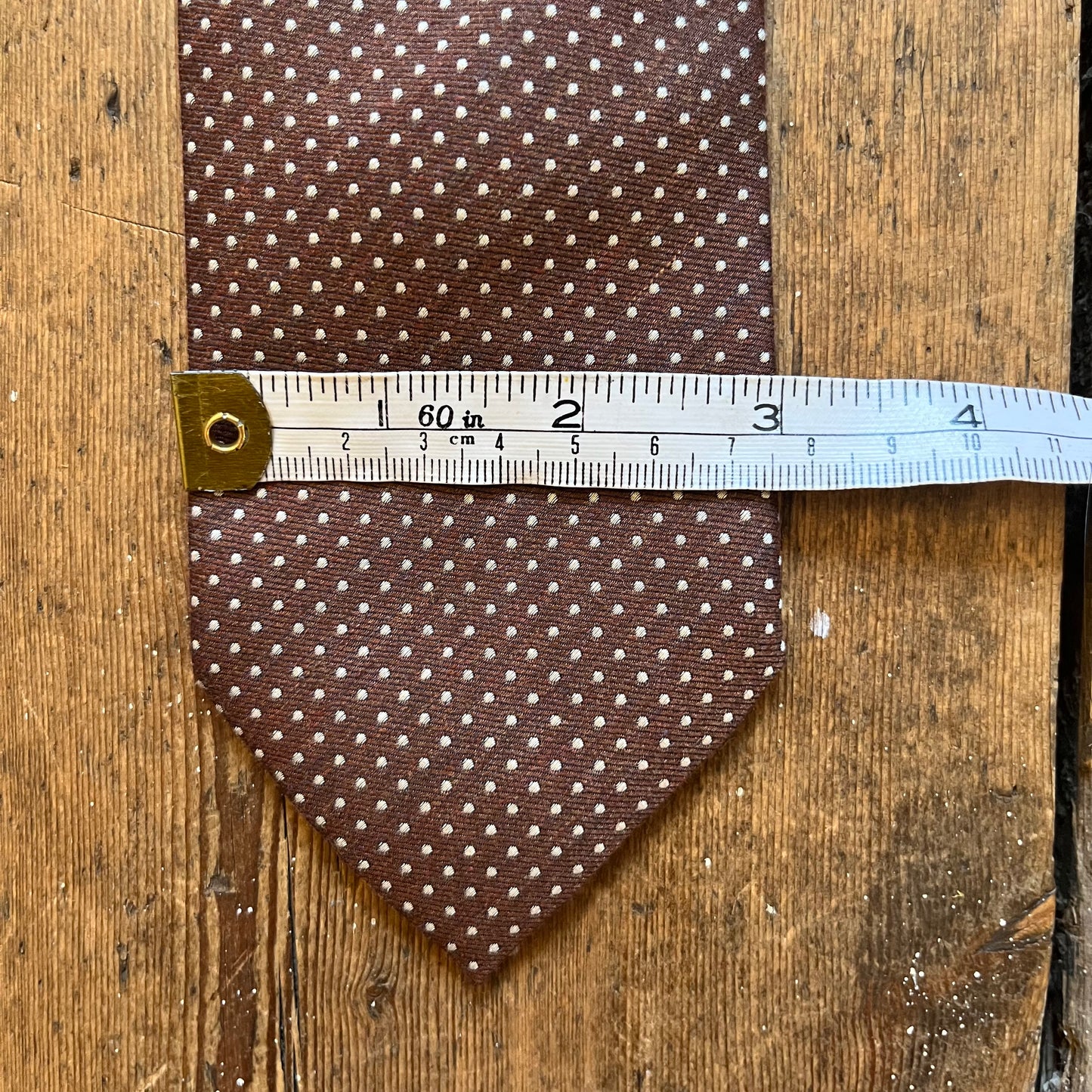 A brown necktie with a small white polka dot pattern is laid flat on a rustic wooden surface, with a metal ruler placed horizontally across the middle to show its width.