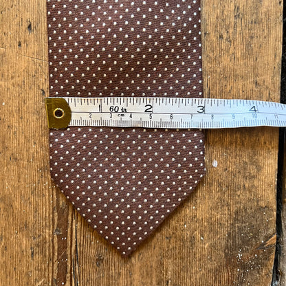 A brown necktie with a small white polka dot pattern is laid flat on a rustic wooden surface, with a metal ruler placed horizontally across the middle to show its width.