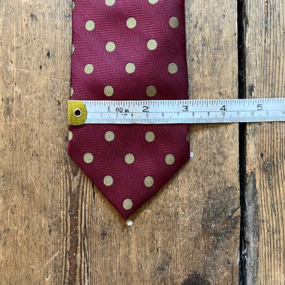 A burgundy necktie with gold polka dots and a subtle herringbone pattern, shown on a rustic wooden background with a metal measuring tape across the middle to indicate size.
