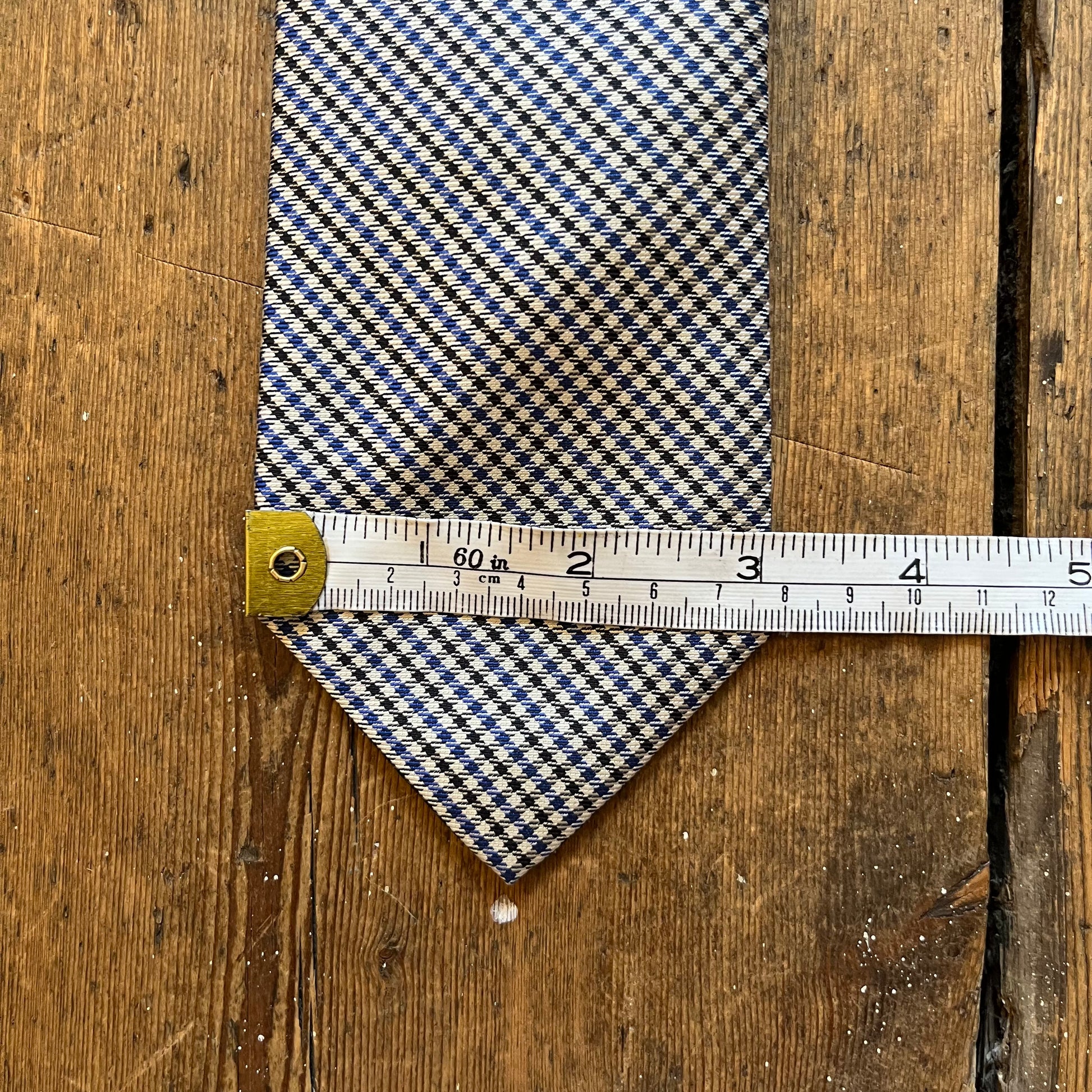 A patterned necktie with a blue, beige, and white design, shown with a metal measuring tape to display its width on a rustic wooden background.