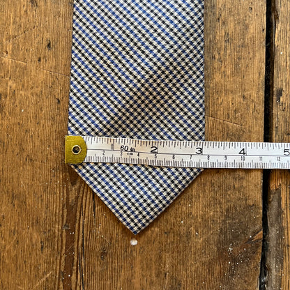 A patterned necktie with a blue, beige, and white design, shown with a metal measuring tape to display its width on a rustic wooden background.