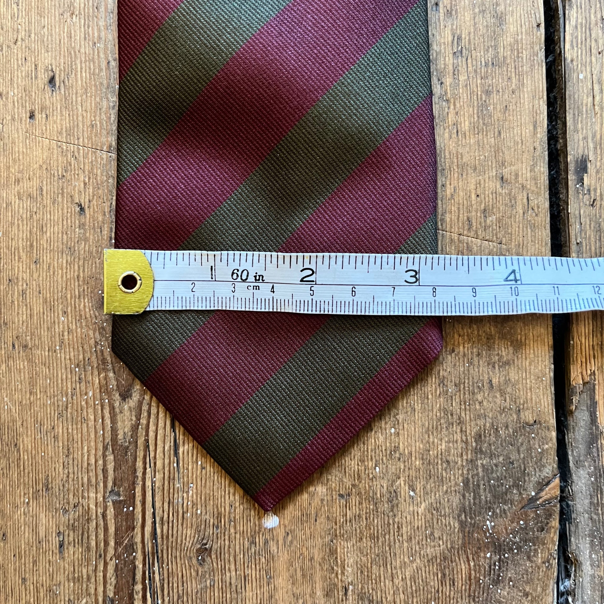 A striped necktie with diagonal bands of burgundy and olive green, shown with a metal ruler laid across the middle to indicate size, on a rustic wooden surface.