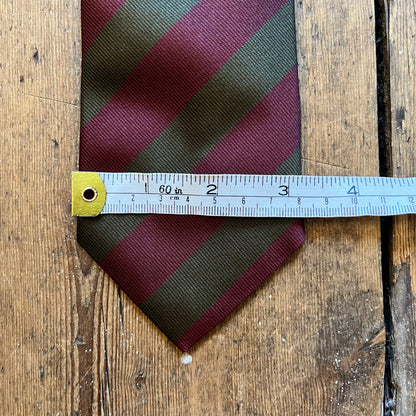 A striped necktie with diagonal bands of burgundy and olive green, shown with a metal ruler laid across the middle to indicate size, on a rustic wooden surface.