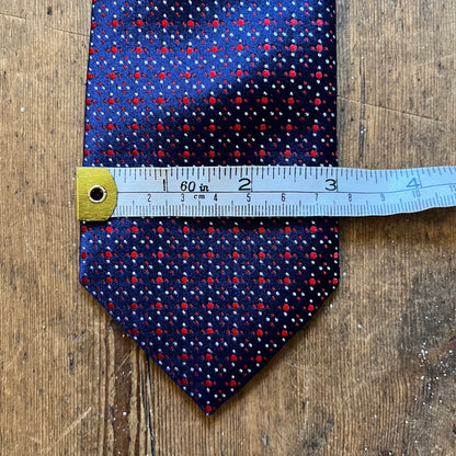 A patterned necktie with a blue base, red and white dots, and a yellow-gold tip, shown on a wooden surface with a measuring tape across the middle for scale.