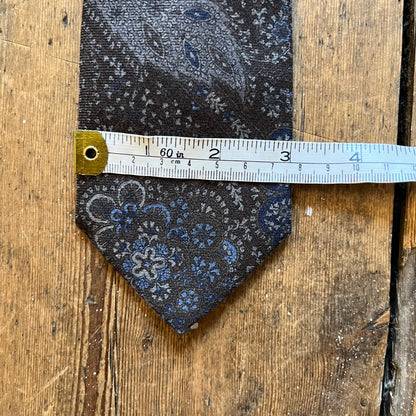 A patterned necktie with a dark charcoal base and blue, gray, and white floral paisley print, shown with a white measuring tape to indicate its size on a rustic wooden background.
