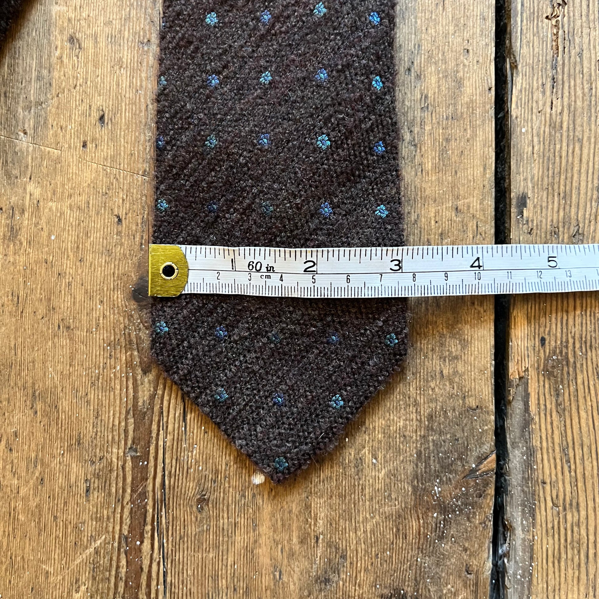 A dark brown textured necktie with small blue and gold dotted pattern, shown flat on a rustic wooden surface with a metal measuring tape laid across the middle to indicate size.