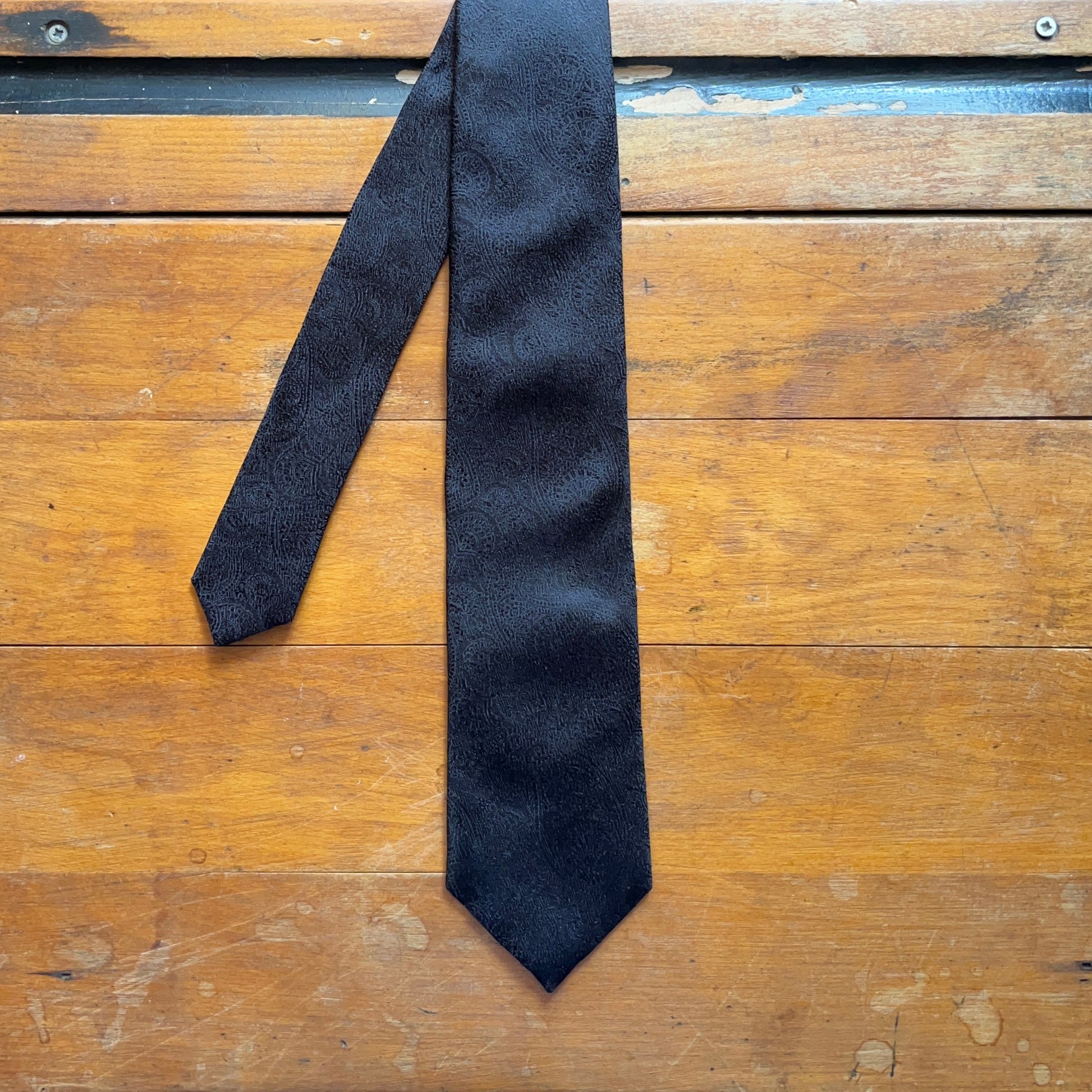 Handmade black silk tie with paisley design set against wooden boards