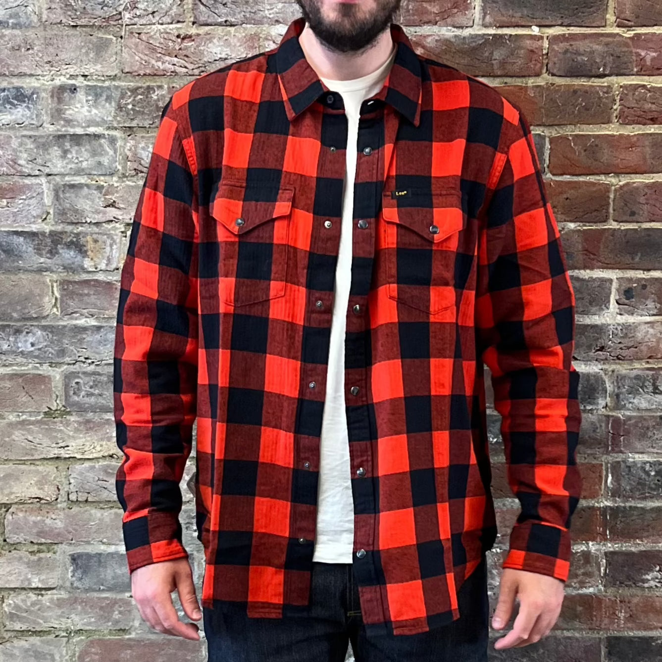 A bearded gentleman standing against a brick wall in a Lee cotton flannel red and black checked shirt over a white tee