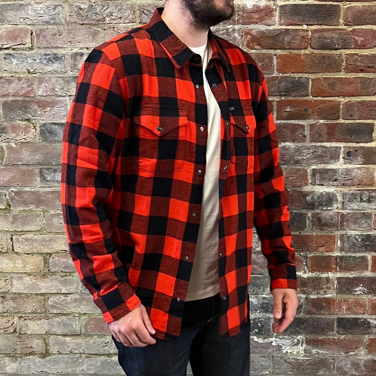 A bearded gentleman standing side on against a brick wall in a Lee cotton flannel red and black checked shirt over a white tee