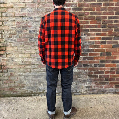 A bearded gentleman standing against a brick wall with his back to the camera in a Lee cotton flannel red and black checked shirt over a white tee