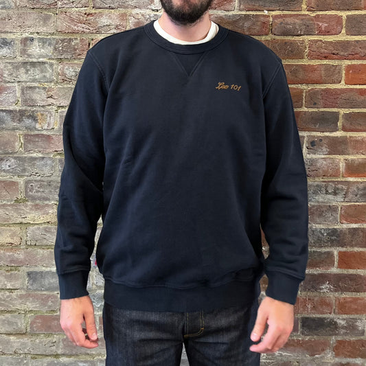 A bearded gentleman standing against a brick wall in a black Lee 101 cotton sweatshirt shirt over a white tee. The sweatshirt has 'Lee 101' embroidered on the chest in brown writing