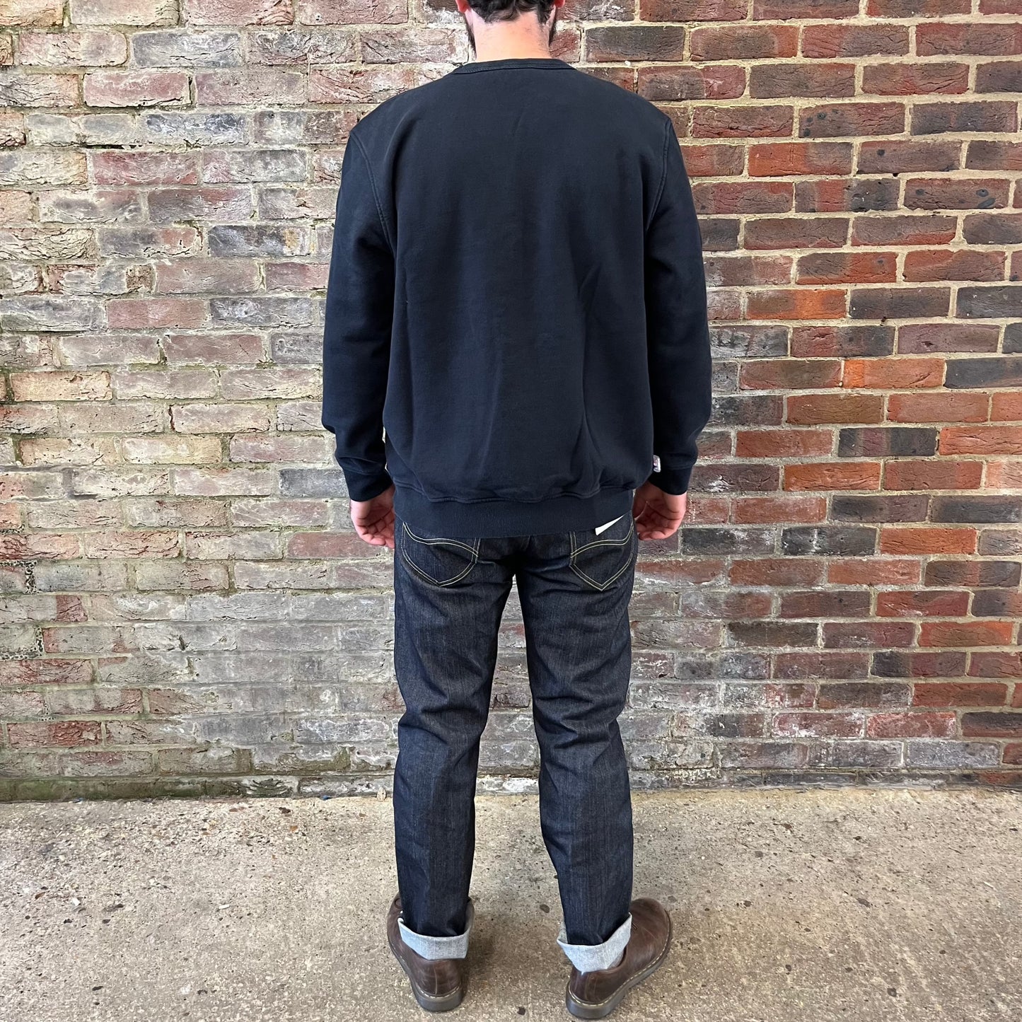 A bearded gentleman facing against a brick wall in a black Lee 101 cotton sweatshirt. He is wearing jeans and Dr. Martens shoes