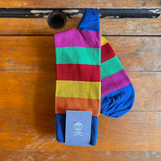 A pair of socks with stripes sit atop a wooden surface. The stripes are Orange, yellow, red, green and pink. The top, heel and toe of the sock are blue. 