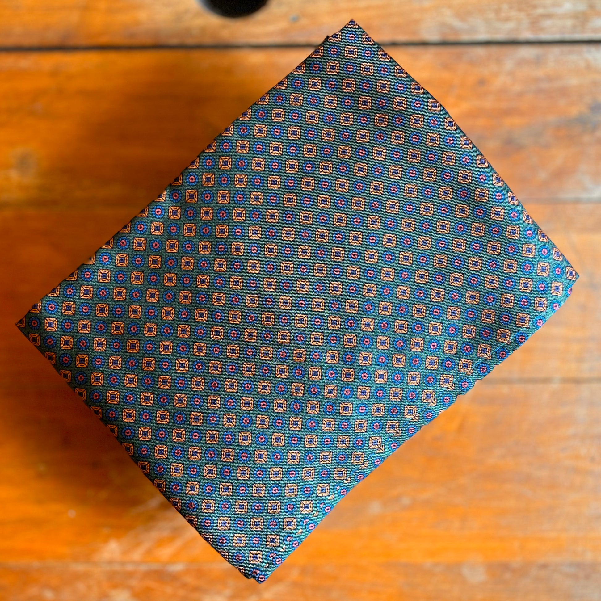 A green tootal scarf with a blue and gold pattern laid on a wooden background