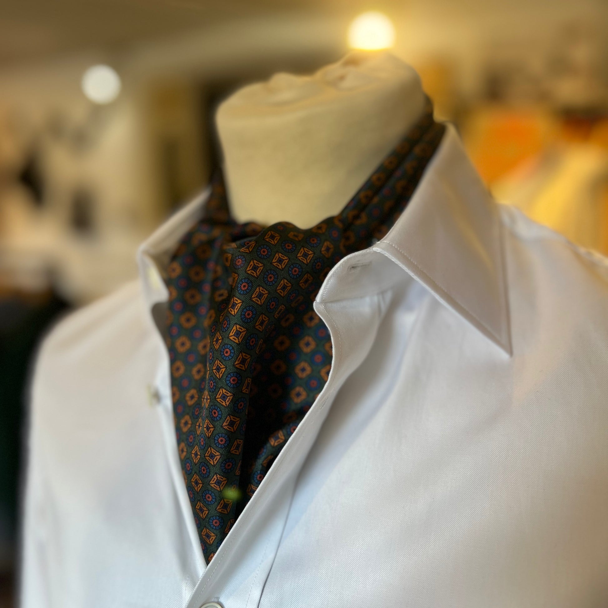 A green tootal scarf with a gold and blue pattern tied in a cravat style on a mannequin