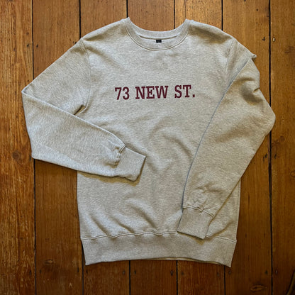 Grey cotton sweatshirt with '73 New Street' in red collegiate writing on the chest, placed on wooden boards