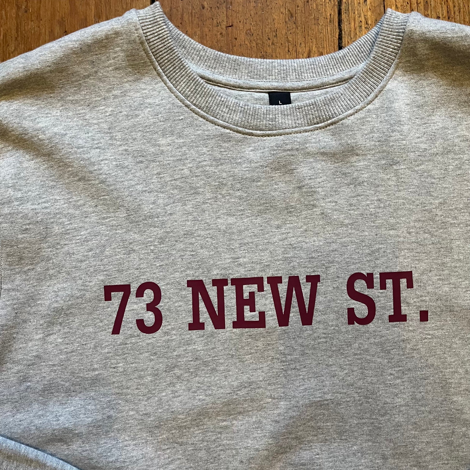 Grey cotton sweatshirt with '73 New Street' in red collegiate writing on the chest, placed on wooden boards