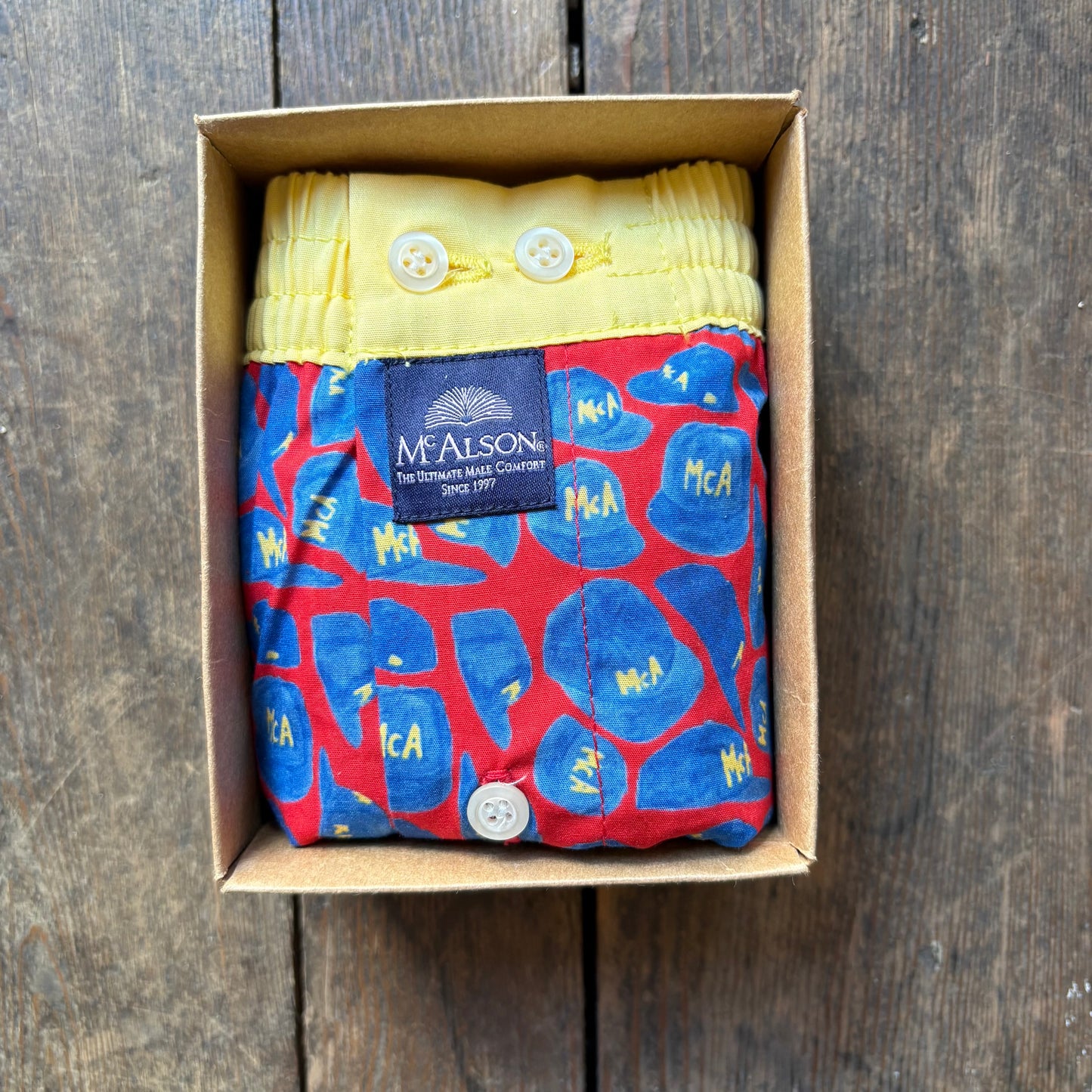 A folded McAlston boxer. The product has a red base with blue abstract shapes and yellow lettering, plus a yellow waistband with white buttons and a dark blue McAlston label.