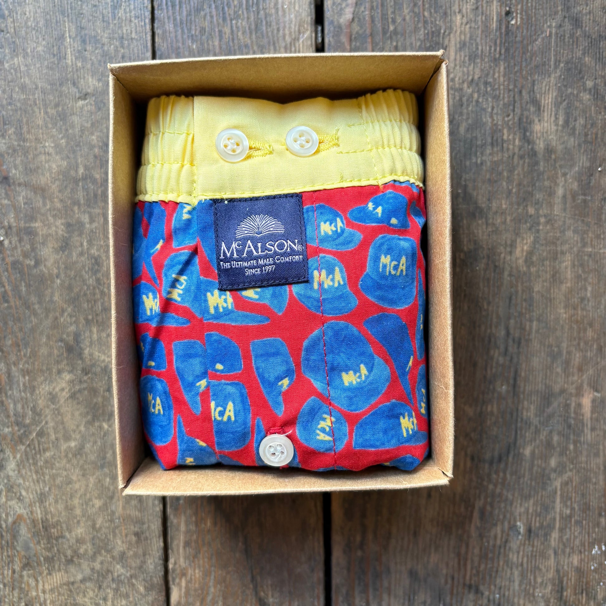 A folded McAlston boxer. The product has a red base with blue abstract shapes and yellow lettering, plus a yellow waistband with white buttons and a dark blue McAlston label.