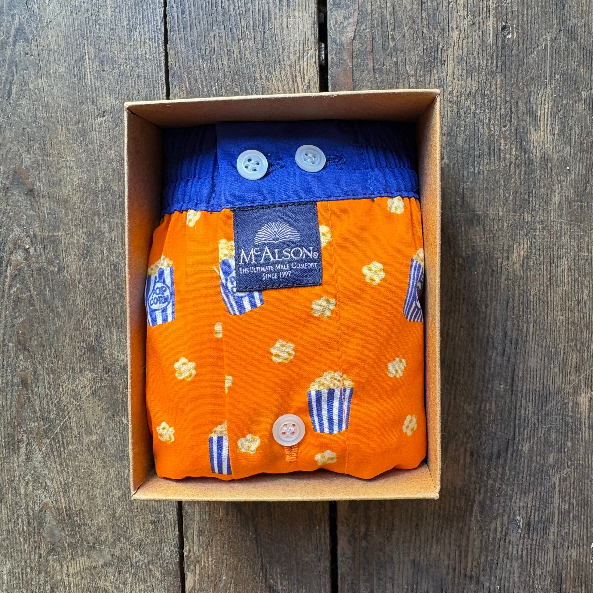 An orange cloth diaper with a popcorn and blue striped pattern, folded inside a brown cardboard box on a dark wooden surface. The diaper has a blue waistband with two white buttons and a visible McAlson brand label.