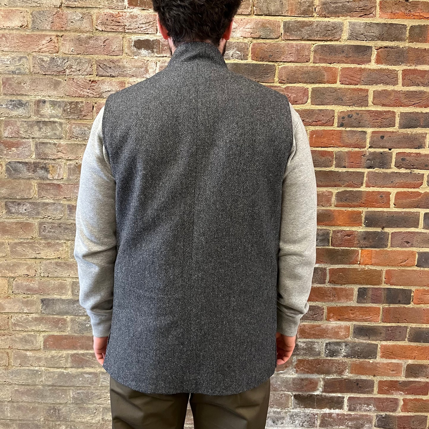 A man stood facing a brick wall wearing a a grey herringbone gilet 