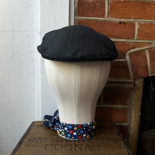 Black pinstripe flat cap on a mannequin head with a polka dot band against a brick wall.