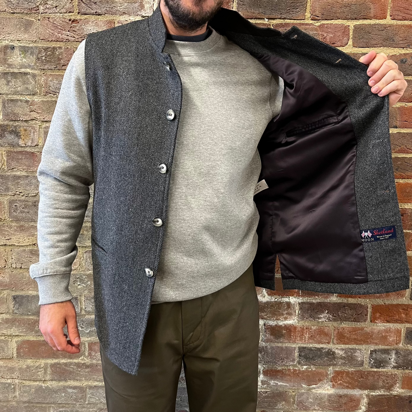 A man stood against a brick wall wearing a a grey herringbone gilet and revealing the purple viscose lining