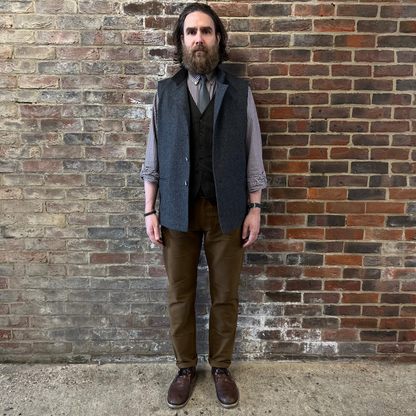 A man stood against a brick wall wearing a a grey herringbone gilet and gingham shirt