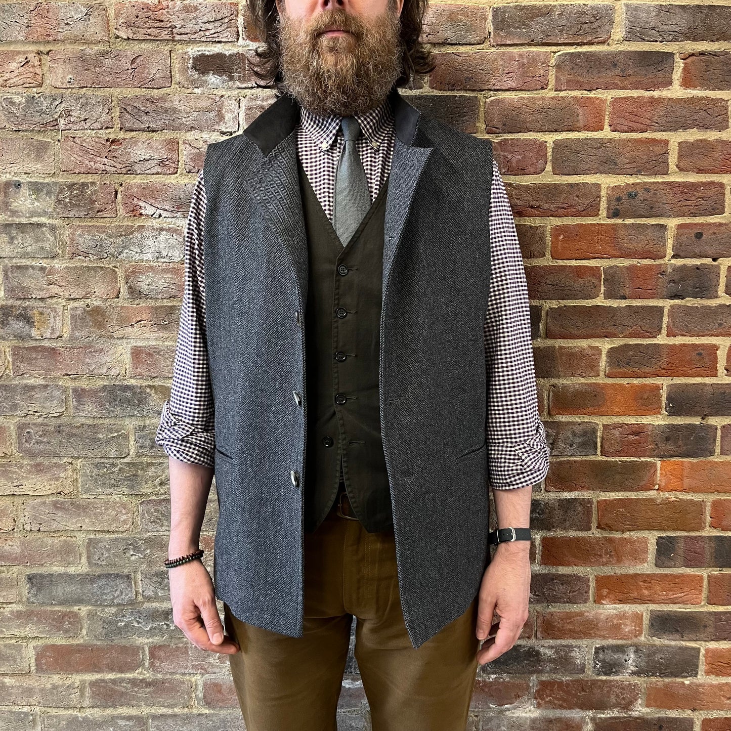 A man stood against a brick wall wearing a a grey herringbone gilet and gingham shirt