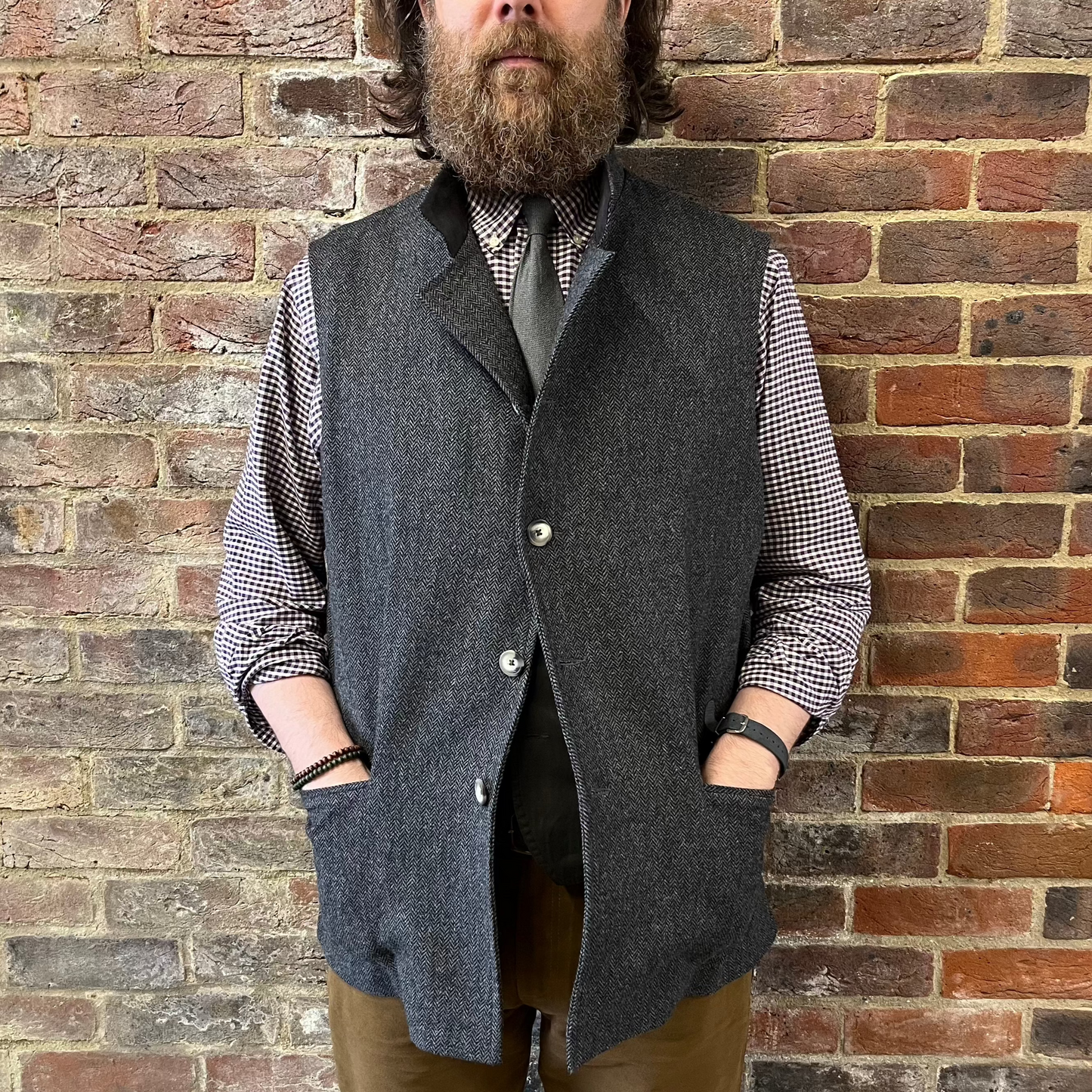 A man stood against a brick wall wearing a a grey herringbone gilet and gingham shirt